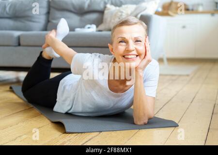 Glückliche lächelnde ältere Frau, die sich streckte und zur Kamera schaute. Bewegung für die körperliche Gesundheit. Wellness für Frauen im Ruhestand. Inländische Yoga-Praxis der flexiblen reifen Frau Ausübung auf Sportmatte Stockfoto