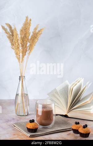 Muffins und ein Glas Kaffee mit Milch auf dem Tisch. Heißes Getränk, Kakao und offenes Buch. Getrocknetes Pampagras in Glasflasche als Vase. Natürliche Pflanzen Dekor o Stockfoto