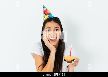Überrascht junge Frau trägt Party-Kegel Hut halten Cupcake mit Kerze, weißem Hintergrund Stockfoto