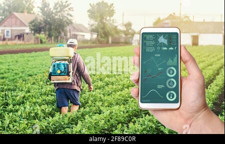 Eine Hand mit einem Telefon auf dem Hintergrund eines Landwirts mit landwirtschaftlicher Rauchnebelsprühmaschine. Pflanzenpflege und Schutz vor Schädlingen und Pilzinfektionen Stockfoto