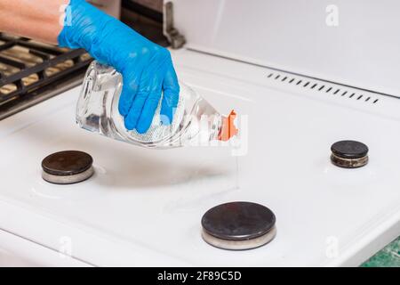 Die Hand der Frau in einem Haushaltshandschuh gießt flüssiges Waschmittel auf einen Gasherd und macht die Hausreinigung der Küche. Stockfoto