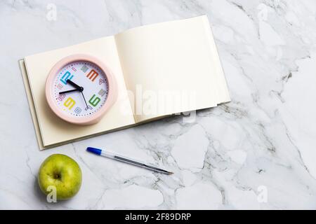 Runder rosafarbener Wecker, quadratisches Notizbuch, Apfel und Stift auf Marmortischoberfläche. Speicherplatz kopieren Stockfoto