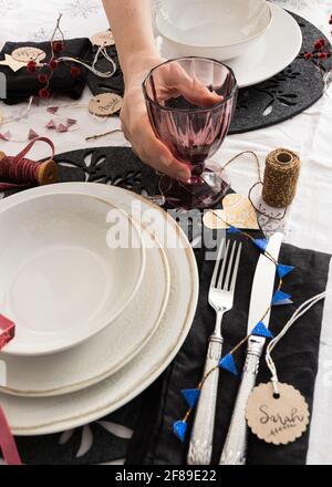 Ein hübsches, transparentes, violettes Glas, das von einer Menschenhand an einem verzierten Weihnachtstisch gehalten wird. Einige verschwommene Namen sind auf runden Tags rund um Th herum zu finden Stockfoto