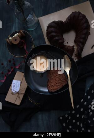 Ein elegantes Dessert auf einem dunklen Tablett, bestehend aus einem schaumig-schäumenden Kaffee und einer Scheibe hausgemachtem Schokoladenkuchen. Ein Geschenk und der ganze Kuchen sind als Hintergrund. Stockfoto