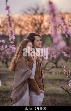 Glückliches Paar in der Liebe bei Sonnenuntergang in blühenden Pfirsichrosengärten. Die junge Frau steht hinter dem Mann, umarmt, küsst mit Zärtlichkeit und Leidenschaft. Romantisches Date, Stockfoto