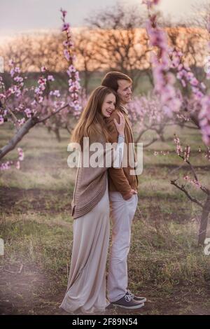 Glückliches Paar in der Liebe bei Sonnenuntergang in blühenden Pfirsichrosengärten. Die junge Frau steht hinter dem Mann, umarmt, küsst mit Zärtlichkeit und Leidenschaft. Romantisches Date, Stockfoto