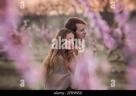 Glückliches Paar in der Liebe bei Sonnenuntergang in blühenden Pfirsichrosengärten. Die junge Frau steht hinter dem Mann, umarmt, küsst mit Zärtlichkeit und Leidenschaft. Romantisches Date, Stockfoto