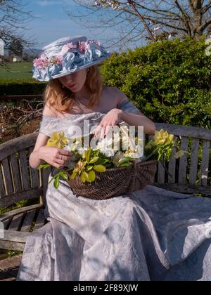 Junge Frau, die auf einer Holzbank sitzt und Blumen in einem Korbkorb umordnet. Nach unten schauen. Tragen Osterhaube und Off-Shoulder-Kleid. Stockfoto