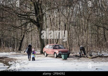 POZNAN, POLEN - 28. Jan 2017: Unbekannte Frau mit Kindern, die an einem geparkten roten Auto vorbeifahren und einen Mann, der an einem kalten Wintertag in einem Park Baumstämme sägt Stockfoto