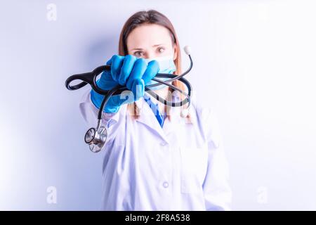 Hübsche junge Ärztin, die ein Stethoskop vor sich hält Maske auf weißem Hintergrund Stockfoto