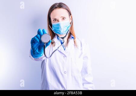 Junge hübsche Ärztin mit Handschuhe Maske und Kleid halten Und ein Stethoskop vorne auf weißem Hintergrund Stockfoto