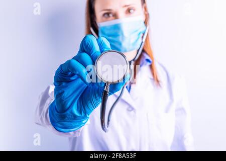 Junge hübsche Ärztin mit Handschuhe Maske und Kleid halten Ein Stethoskop vorne auf weißem Hintergrund Stockfoto