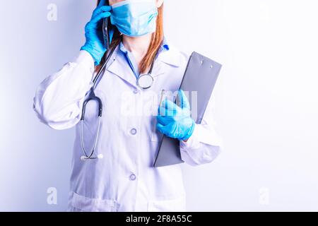 Kaukasische junge Frau trägt Stethoskop Kleid und Handschuhe und Maske hält Handy an einem Ohr und Berichte im anderen. Technologie und Medizin c Stockfoto