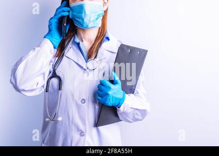 Kaukasische junge Frau trägt Stethoskop Kleid und Handschuhe und Maske hält Handy an einem Ohr und Berichte im anderen. Technologie und Medizin c Stockfoto