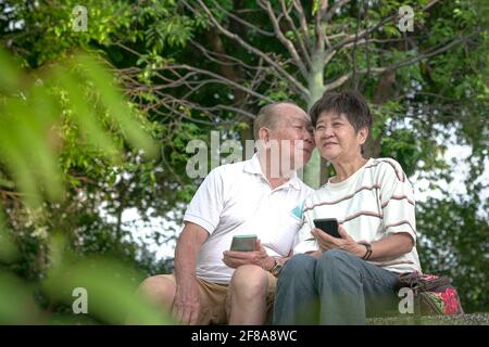 Älterer asiatischer Mann küsst die Wange seiner Frau. Außenumgebung. Liebevolles Beziehungskonzept. Stockfoto