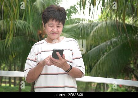 Ältere Erwachsene asiatische Frau lächelt, während sie im Park ihr Telefon liest. Stockfoto