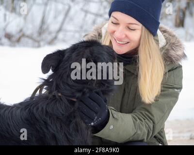 Junge Frau spielt mit lustigen schwarzen Hund mit Schnee. Auf dem Hintergrund. Spielen mit dem Schnee. Liebenswert Hund genießen ihre Zeit, Winter Zeit. Speicherplatz kopieren Stockfoto
