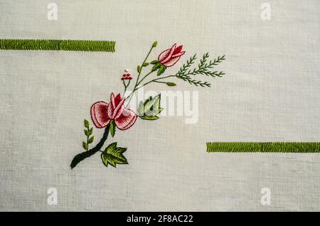 Zwischen grünen Bändern bestickt, ein Zweig mit roten Rosen und einer Knospe, verflochten mit einem Nadelzweig und roten Beeren auf einem weißen Baumwollstoff Stockfoto