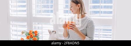 Glückliche Frau mittleren Alters, die Kaffee trinkt und Katze anschaut. Junge Frau mit kurzen Haaren, die Tee aus der Tasse trinkt und mit dem Haustier spielt. Entspannen und Freizeit t Stockfoto