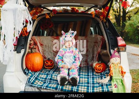 Trick or Trunk. Traurig verärgert Baby im Einhorn Kostüm feiert Halloween im Kofferraum eines Autos. Niedliches Kleinkind feiert Oktoberfest im Freien. Soziale di Stockfoto