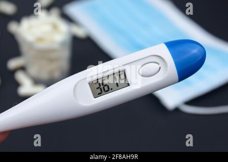 Hand hält weißes elektronisches Thermometer im Hintergrund mit Packungen von Tabletten und blauer medizinischer Gesichtsmaske. Stockfoto
