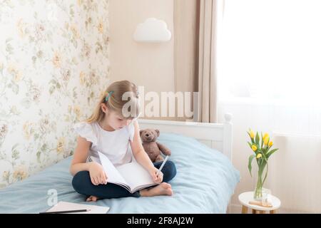 Ein kleines Mädchen sitzt auf dem Bett im stilvollen Schlafzimmer und schreibt in einem blauen Buch. Bildungskonzept. Zu Hause Schulbildung. Hausaufgaben. Gelbe Tulpen auf dem Tisch Stockfoto