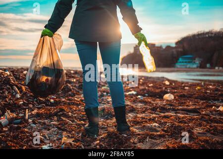 Tag der Erde. Eine Freiwillige steht an einem schlammigen Strand mit einem Sack Müll. Rückansicht. Stockfoto