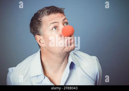 Mann mittleren Alters mit blauem Hemd und roter Clownsnase Posiert vor blauem Hintergrund im Studio Stockfoto