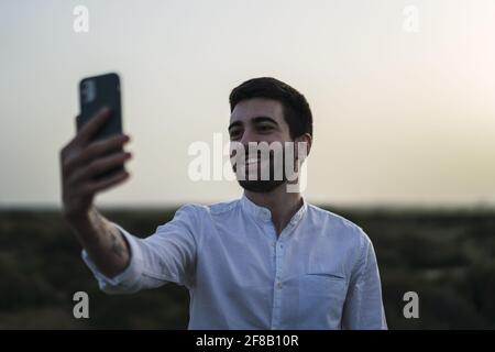 Flacher Fokus eines jungen kaukasischen Mannes, der einen weißen Mann trägt shirt und ein Selfie auf einem Feld Stockfoto