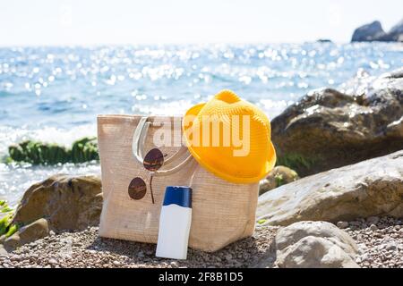 Strandtasche mit einem weißen Sonnenschutzrohr an einem Kiesstrand in der Nähe des Meeres. Reisen, Strandurlaub im Resort, Gläser, gelber Hut. Schutz der s Stockfoto