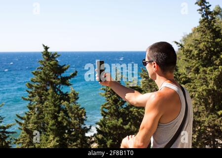 Männlicher Tourist macht Fotos auf einem Smartphone von schönen Blick auf das Meer in einer bergigen Gegend von der Spitze unter Nadelbäumen. Videokommunikation, usi Stockfoto