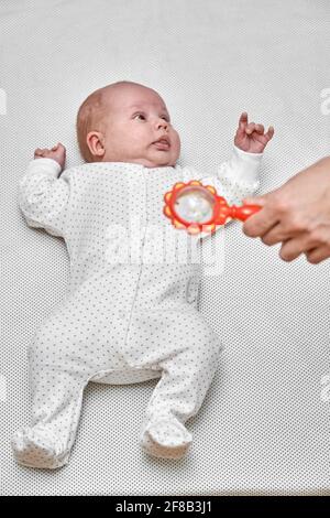 Süßes kleines Neugeborenes mit winzigen Fingern, die weißen Körper tragen Lügt und schaut auf die Mutter, die orangefarbene Maraca in der Nähe hält Stockfoto