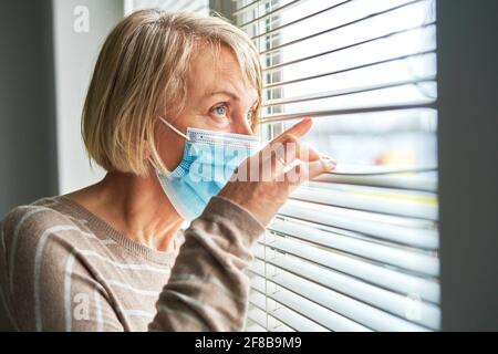 Eine unter Quarantäne gestellte ältere Frau mit Maske schaut durch die Jalousien Stockfoto