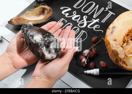 Nahaufnahme einer weiblichen Hand, die eine schimmelige Avocado auf einer Tafel mit Schriftzügen und verfaulten Früchten hält. Selektiver Fokus. Konzept der Stop wa Stockfoto