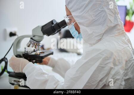 Mikrobiologe in einem Hazmat-Anzug, der ein mikroskopisches Exemplar beobachtet Stockfoto