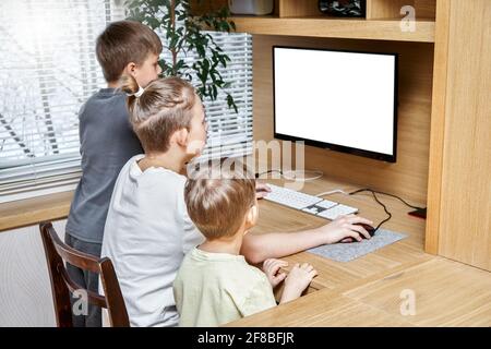Entzückende Kinder sitzen auf einem Stuhl neben dem braunen hölzernen Computertisch Blick in die weiße Anzeige am Fenster auf der Rückseite des Hauses Stockfoto