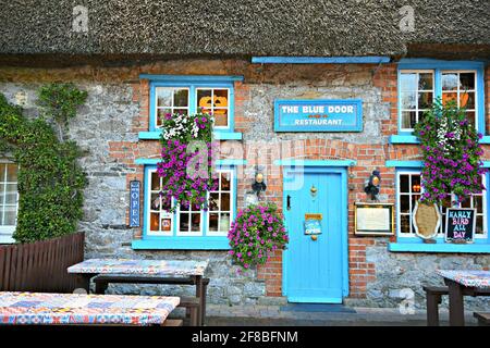 Blick von der Fassade auf das Blue Door Pub and Restaurant mit der blauen Holztür und den Fenstern, den Backsteinwänden und dem Strohdach in Adare Limerick Irland. Stockfoto