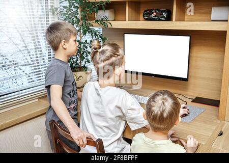 Entzückende Kinder sitzen auf einem Stuhl neben dem braunen hölzernen Computertisch Blick in die weiße Anzeige am Fenster auf der Rückseite des Hauses Stockfoto