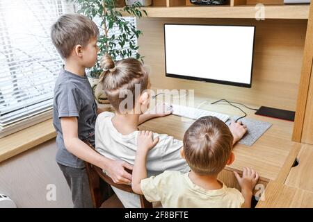 Entzückende Kinder sitzen auf einem Stuhl neben dem braunen hölzernen Computertisch Blick in die weiße Anzeige am Fenster auf der Rückseite des Hauses Stockfoto