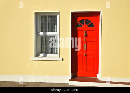 Ländliche Hausfassade mit einer roten Holztür, einem Bronzestiel und einem Briefkasten und einem weißen Fenster an einer gelb gestrichenen Wand in Adare County Limerick Irland Stockfoto