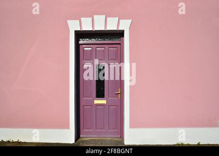 Ländliche Hausfassade mit einer blassrosa Wand und einer Pflaume-Eingangstür mit weißen Zierleisten im malerischen Dorf Adare in der Grafschaft Limerick, Irland. Stockfoto