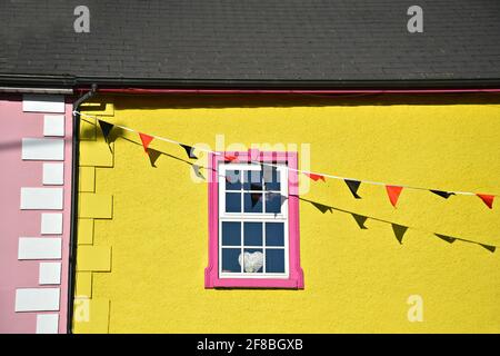 Lokale Gebäudefassade mit einem weißen Holzfenster, rosa Zierleisten und einer leuchtend gelb gestrichenen Wand mit bunten Papierfahnen in Adare, Limerick, Irland. Stockfoto