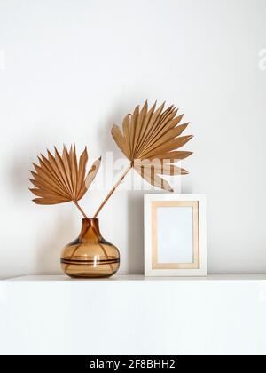 Leerer weißer Fotorahmen neben einer Glasvase mit getrockneten Palmblättern. Stockfoto
