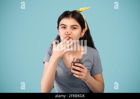 Jugendliches Indial-Mädchen, das Schokoladencreme aus dem Glas isst, sich erfreut fühlt und ihren Finger auf blauem Studiohintergrund mag Stockfoto