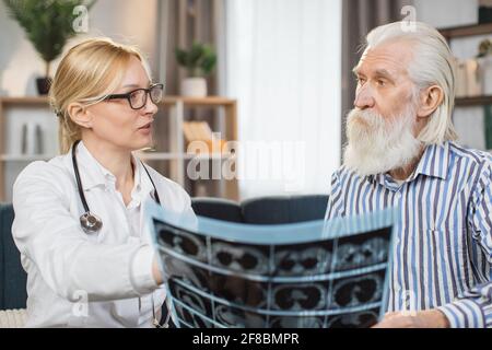 Nahaufnahme des Porträts einer attraktiven Ärztin im Alter von 40 Jahren, die zusammen mit einem älteren bärtigen männlichen Patienten über die Ergebnisse des Röntgenbildes spricht, während er ihn zu Hause besucht. Stockfoto