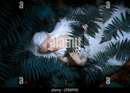 Foto eines ätherischen geheimnisvollen Elfmädchen mit silbernen langen Haaren in einem schönen weißen Kleid und einer silbernen Tiara, die in der Nähe des Baumes in den Farnen liegt. Stockfoto