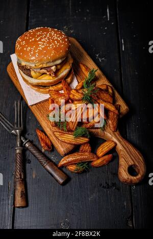 Gebratene Kartoffelkeile mit Kräutern und köstlichem Doppel-Burger auf einem hölzernen Schneidebrett. Rustikaler dunkler Hintergrund. Fastfood-Fotografie. Vertikale Aufnahme Stockfoto