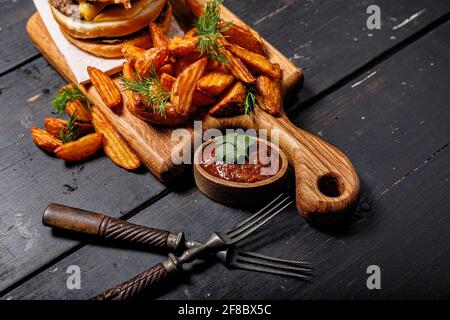 Appetitlich gebratene Kartoffelkeile und Burger auf einem hölzernen Schneidebrett. Große Portion gebackene Kartoffeln mit Kräutern und Tomatensauce. Fast Food foodporn Stockfoto