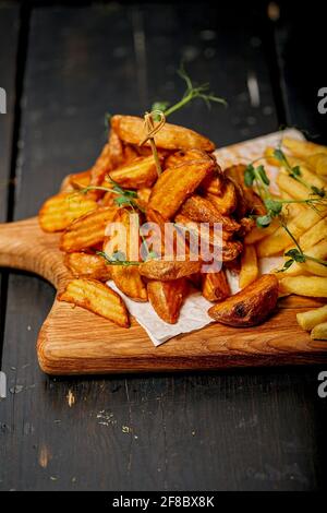 Nahaufnahme von frittierten Kartoffelkeilen auf einem Holzschneidebrett. Große Portion gebackene Kartoffeln mit Sauce und Kräutern. Hausgemachte pommes frites mit Kartoffelscheibe aus biologischem Anbau Stockfoto