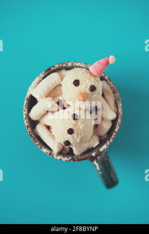 Schneemann aus Kokosnussbällen in weihnachtsmütze, der in einer Tasse mit heißer Schokolade und Marschmalow auf blauem Hintergrund liegt. Weichfokus. Weihnachtsfeier kontra Stockfoto